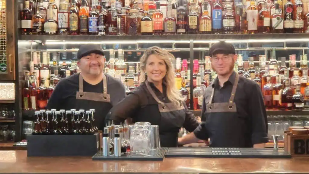 three bartenders behind a bar with a huge wall of bourbons and whiskeys 