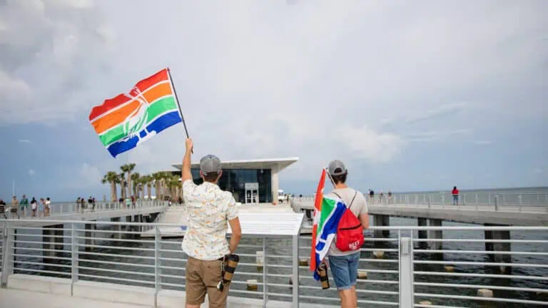 two people hold a multi color flag in the wind on a waterfront pier