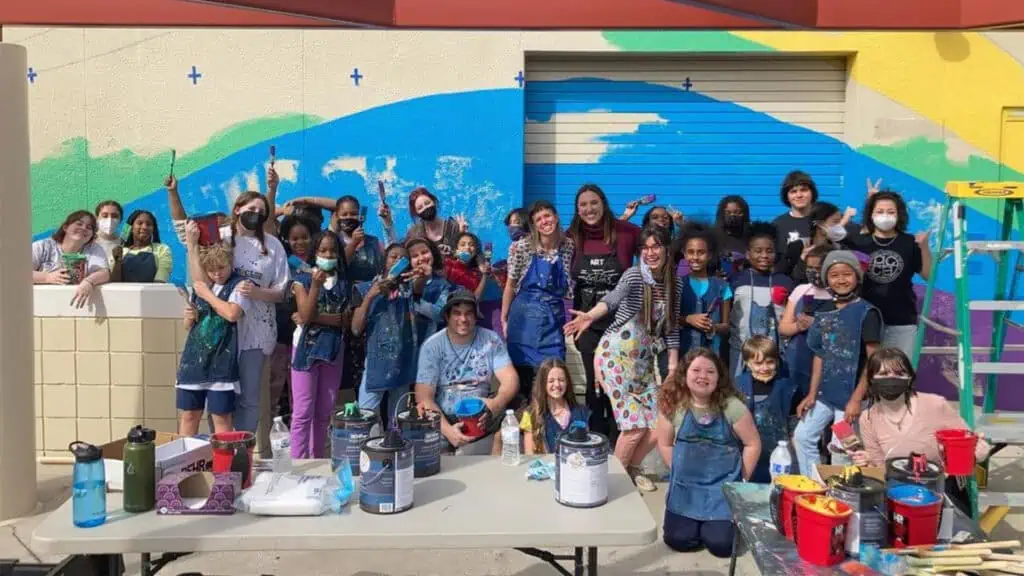 a group kids pose in front of a colorful mural they painted 