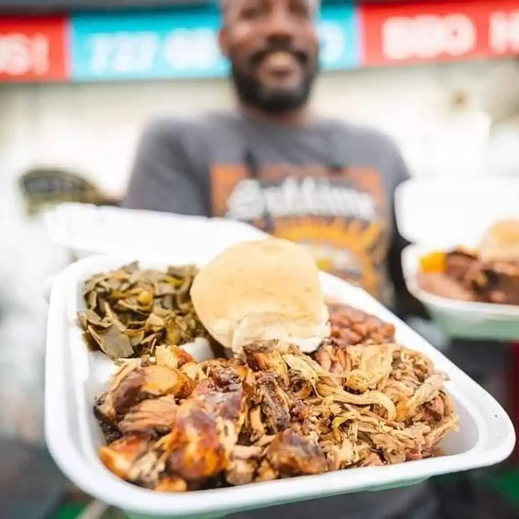 a man holds a plate of pulled pork with traditional BBQ sides in a white to-go container 