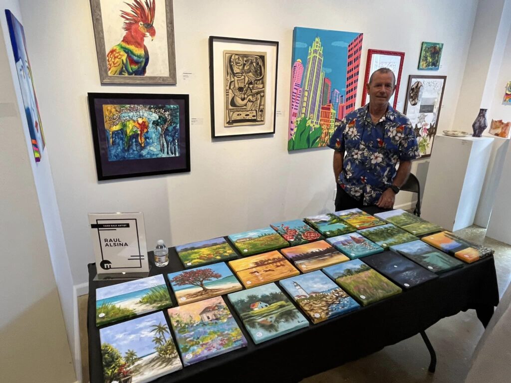 man standing behind table covered in paintings for sale.