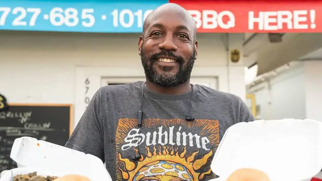 a man holds two to-go containers outside of a small BBQ restaurant.