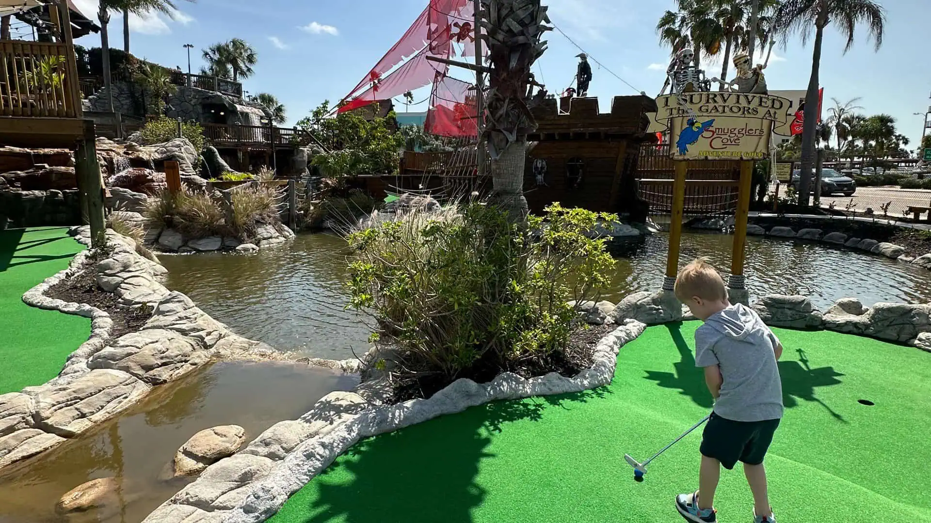 small boy playing mini golf on astro turf near a pond