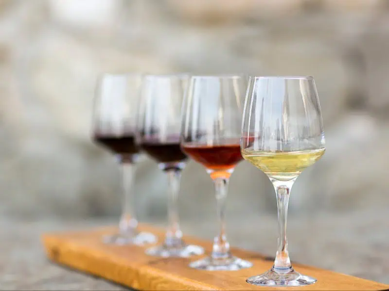 A wooden board holds a flight of four wine glasses, each filled with different varieties of wine ranging from white to deep red.