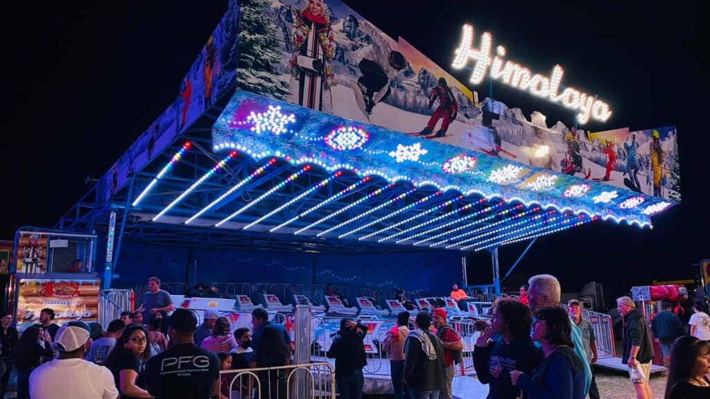 People gather around the brightly lit Himalaya amusement ride at night, waiting for their turn.