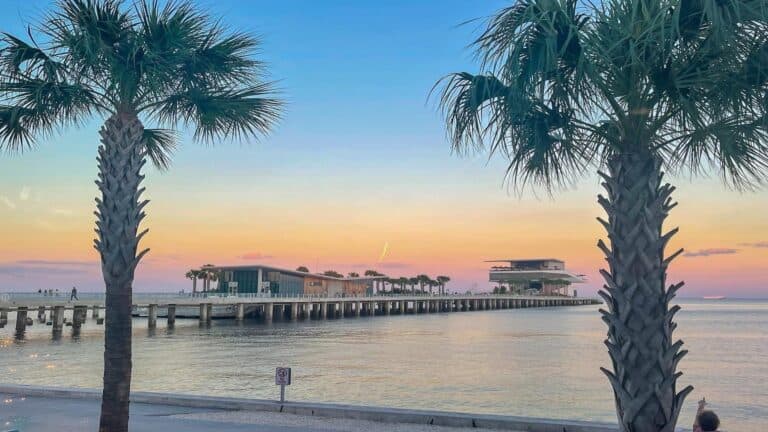 The St. Pete Pier at sunset, framed by two palm trees with a pastel-colored sky in the background.