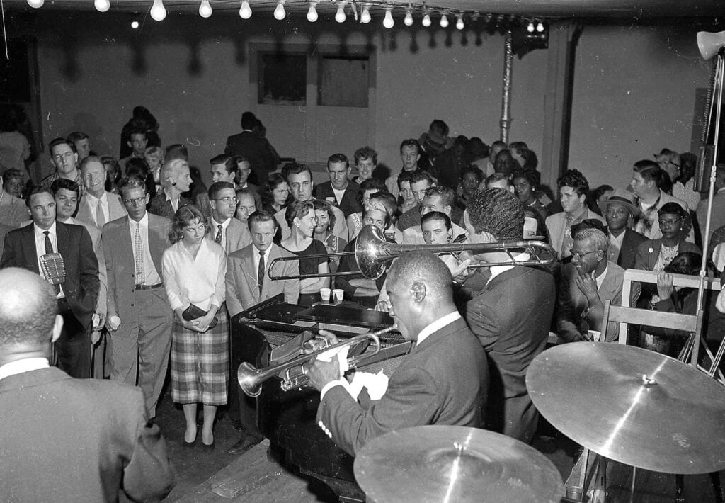 An audience watches a jazz band perform in an intimate, crowded venue.