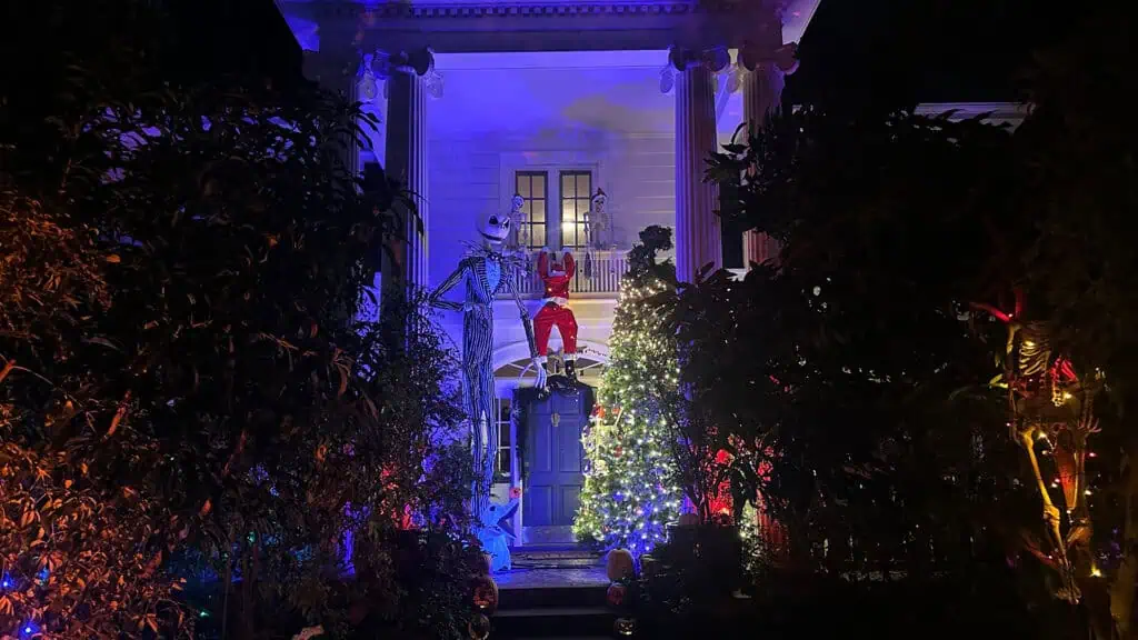 A spooky Halloween display features a Jack Skellington on the porch of a decorated house, illuminated with colorful lights and surrounded by trees and festive ornaments.