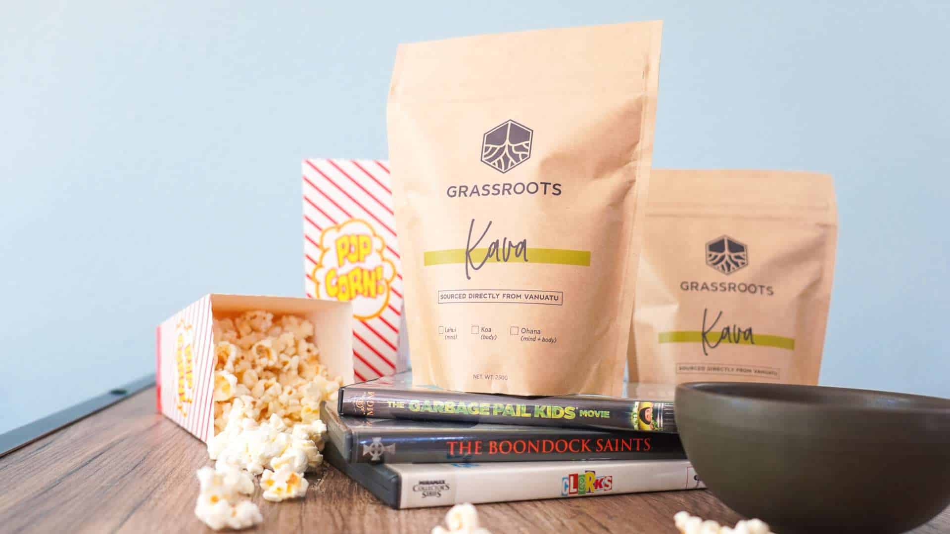 A cozy setup with a bag of Grassroots Kava, a popcorn container spilling popcorn, a bowl, and DVDs on a wooden surface.