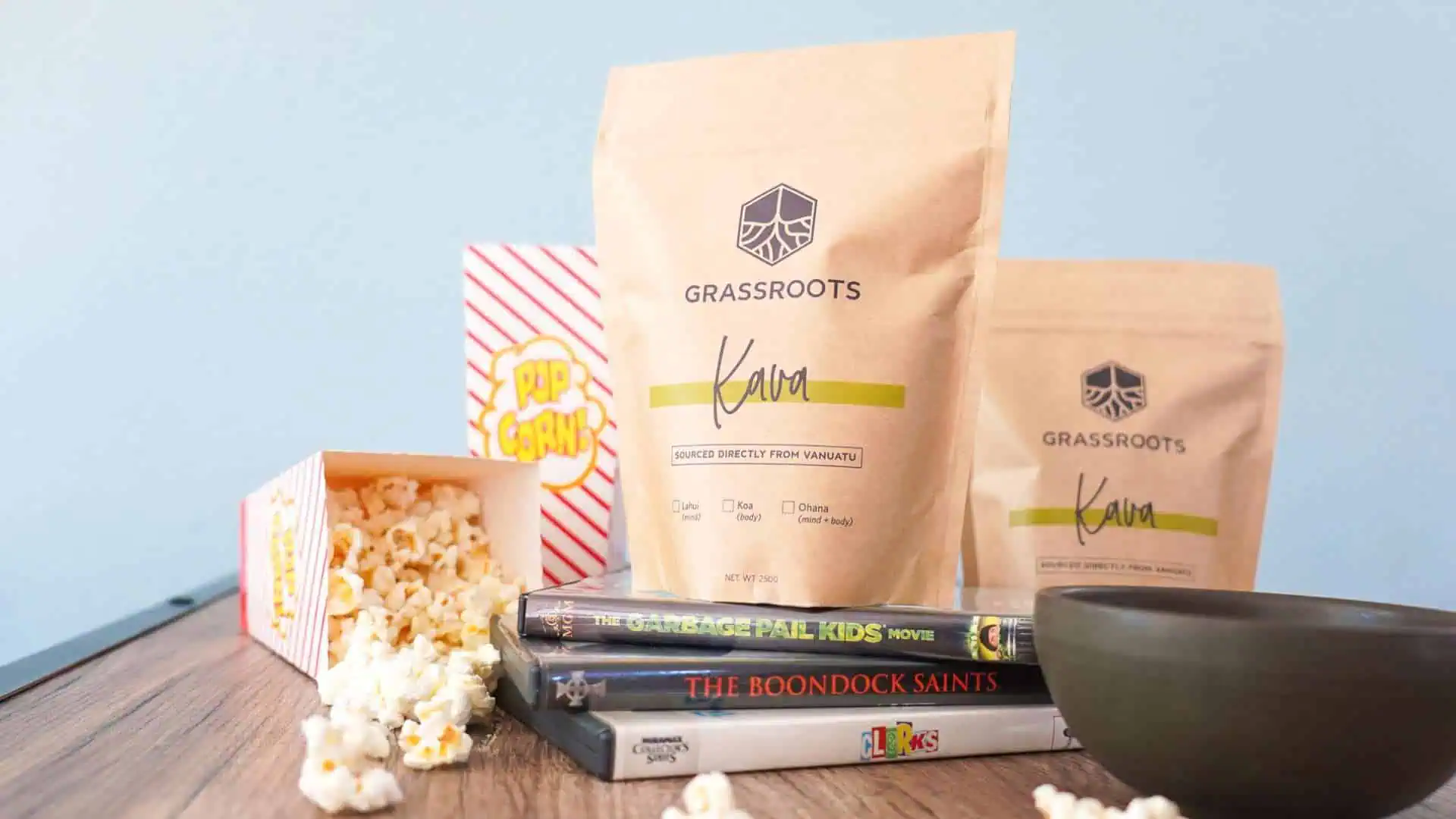 A cozy setup with a bag of Grassroots Kava, a popcorn container spilling popcorn, a bowl, and DVDs on a wooden surface.