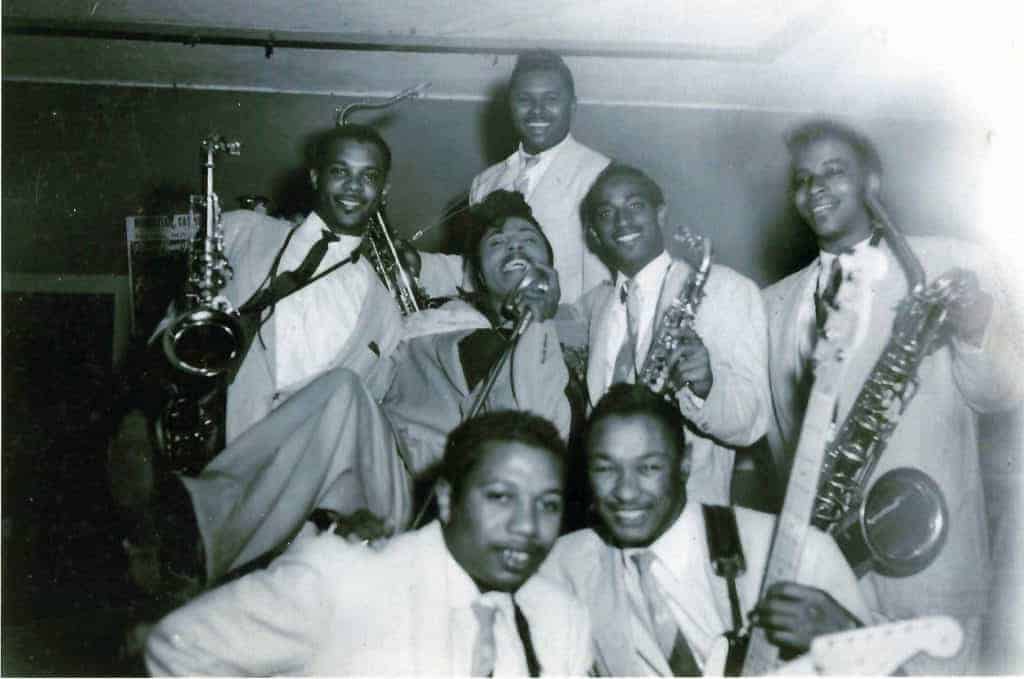 Smiling jazz musicians gather closely for a group photo, holding their instruments.