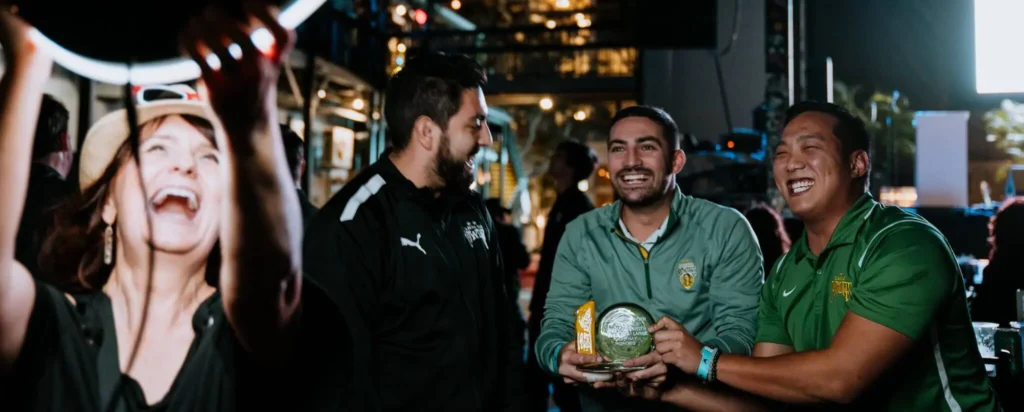 A group of people laugh and celebrate, with one person holding a trophy in a lively nighttime setting.