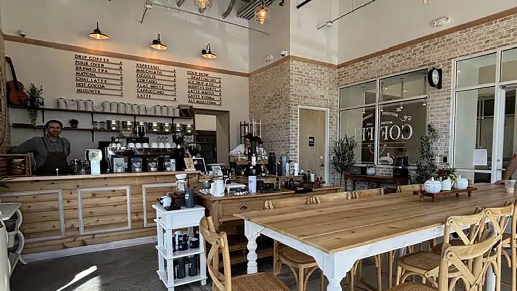 A cozy, rustic coffee shop interior with a large wooden communal table, menu boards on the wall, and various coffee-making equipment behind the counter.