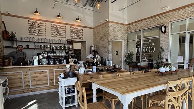 A cozy, rustic coffee shop interior with a large wooden communal table, menu boards on the wall, and various coffee-making equipment behind the counter.