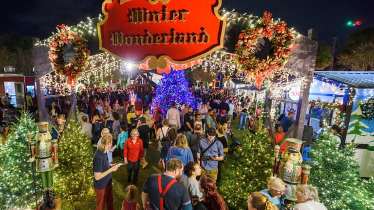 A festive outdoor "Winter Wonderland" event at night, filled with holiday lights, decorated Christmas trees, wreaths, and a large crowd enjoying the holiday atmosphere.