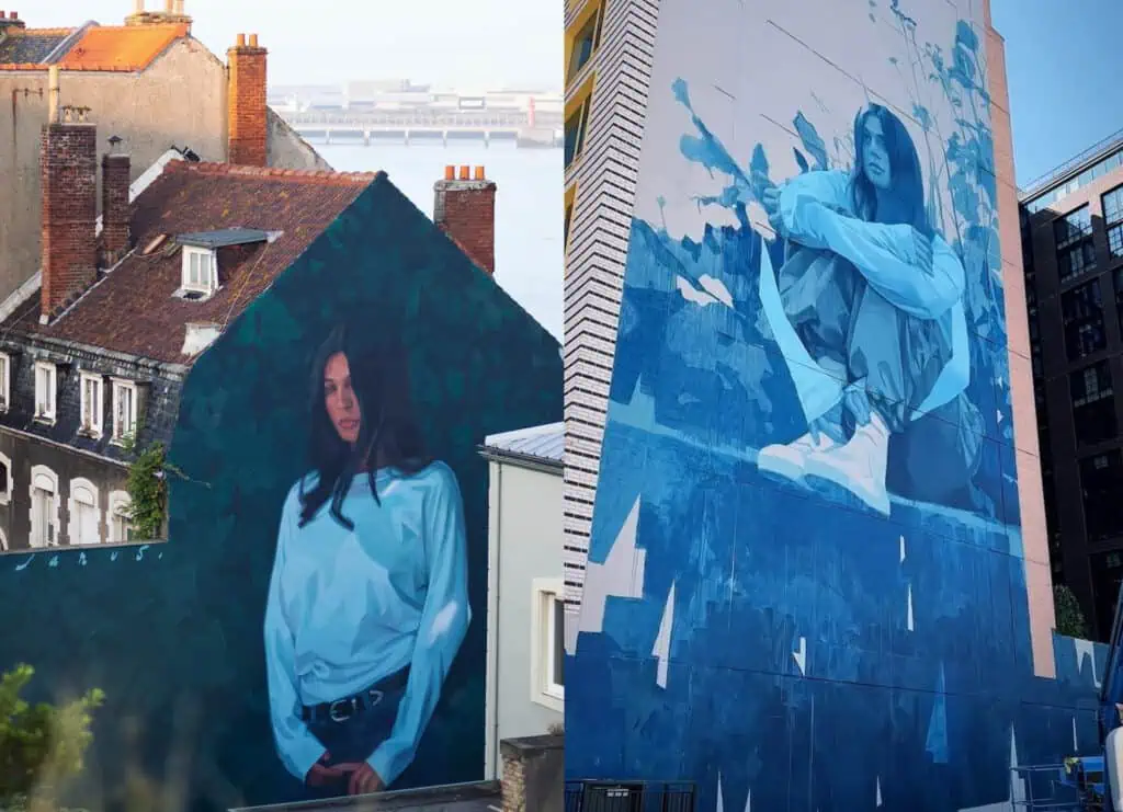 Two large murals painted on building walls depict women in shades of blue, one showing a woman standing against a dark background and the other showing a woman seated with her arms wrapped around her knees.