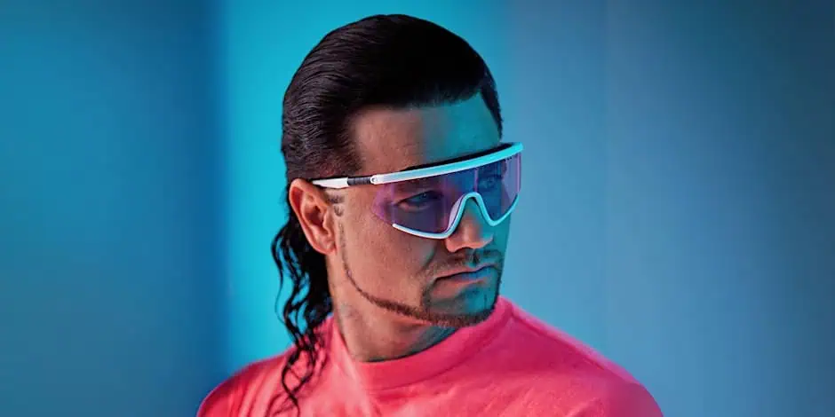 A man in a bright pink shirt and futuristic sunglasses with a mullet hairstyle gazes off to the side against a blue background.