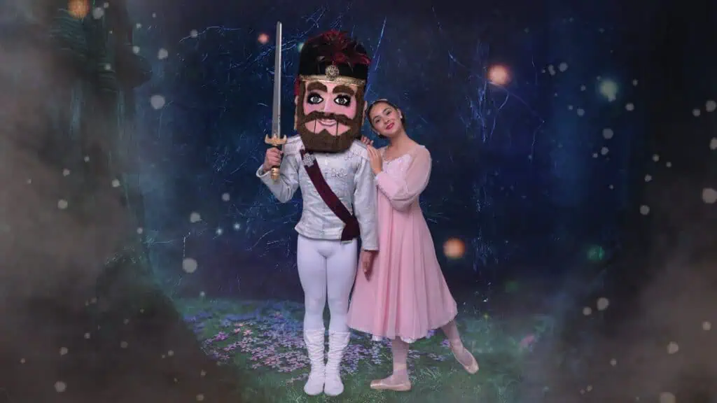 A Nutcracker character with a large, detailed mask and a sword stands beside a ballerina in a pink dress in an enchanted forest setting.