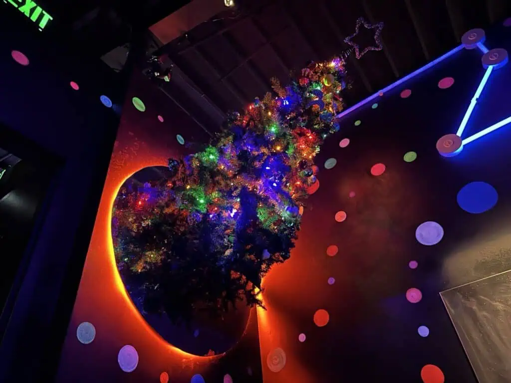 An upside-down Christmas tree adorned with colorful lights, set against a vibrant, neon-lit wall decorated with glowing polka dots and a star at the top.