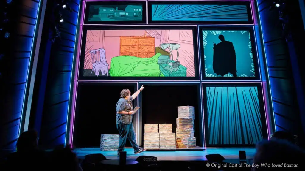 A performer on stage points at large comic-style panels projected above, with stacks of paper props nearby, in a theatrical setting for The Boy Who Loved Batman.