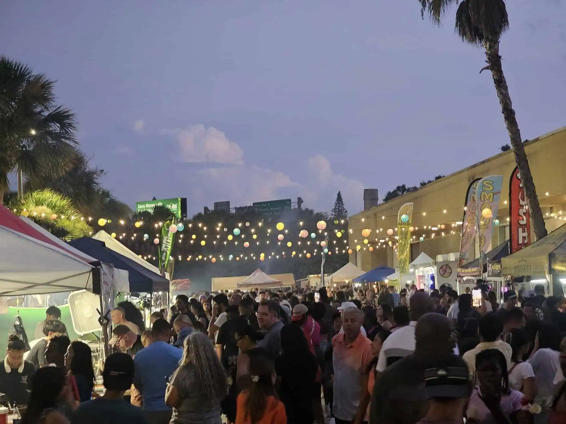 A bustling outdoor night market is packed with people, food stalls, and colorful string lights hanging above the crowd.