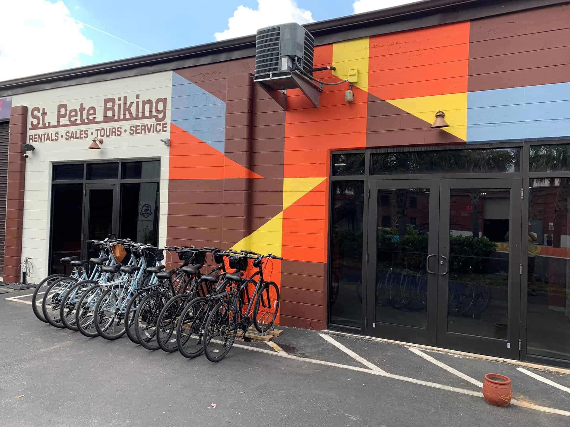 A storefront with the sign "St. Pete Biking" above the entrance, featuring a lineup of bicycles parked in front of a colorful mural.