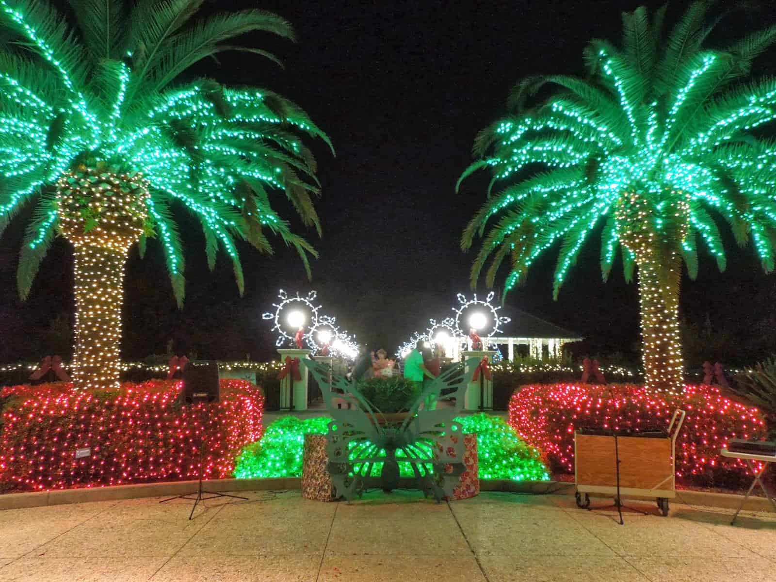 Palm trees wrapped in glowing green and gold lights stand above festive red and green light displays, with decorative snowflakes and a lit gazebo in the background.