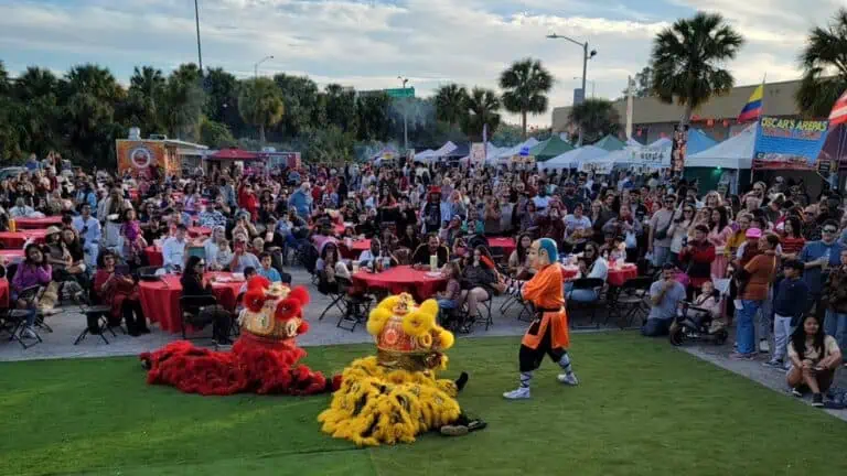 a group of people gathered outside at night for a big Lao Street Food Night Market. Perofrmers are dressed as dragons and perform on a green turf area.