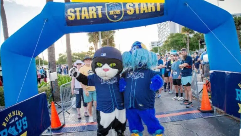 Rays mascots, DJ Kitty and Raymond, stand under the inflatable "Start" arch at a community 5K run event, with participants gathered in the background.