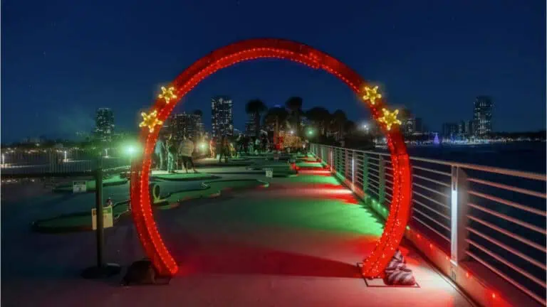 A brightly lit holiday-themed arch with glowing stars overlooks a miniature golf course at night, with city lights twinkling in the background.