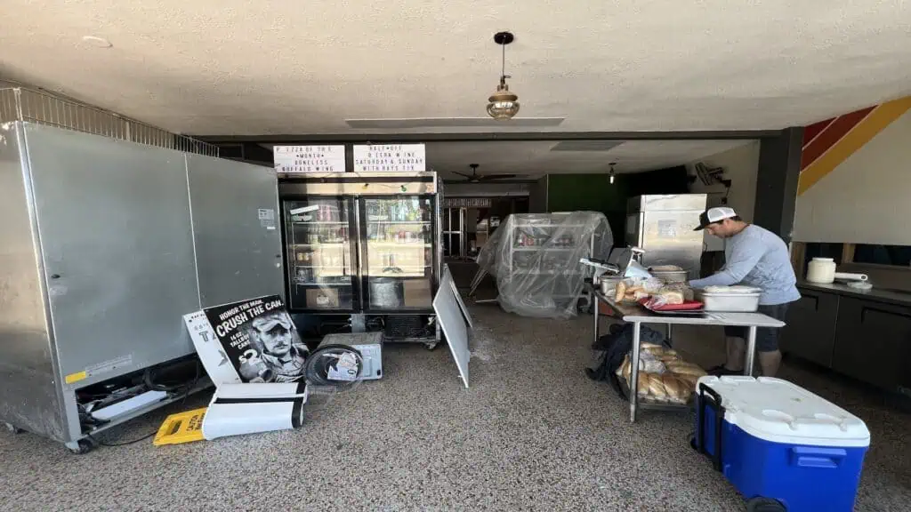 A man prepares food at a worktable surrounded by refrigerators, storage equipment, and signs in a partially set-up restaurant space.