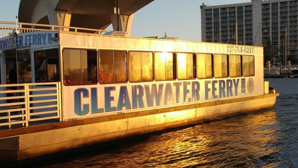 a ferry vessel at sunset on the water. A white and blue banner is draped over the side. The ferry is docked under a tall bridge.