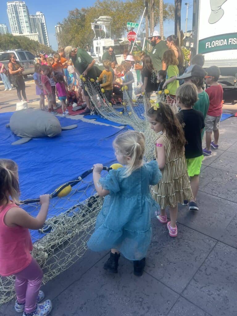 A group of children helping to hold a large fishing net during an educational event with Florida Fish and Wildlife staff.