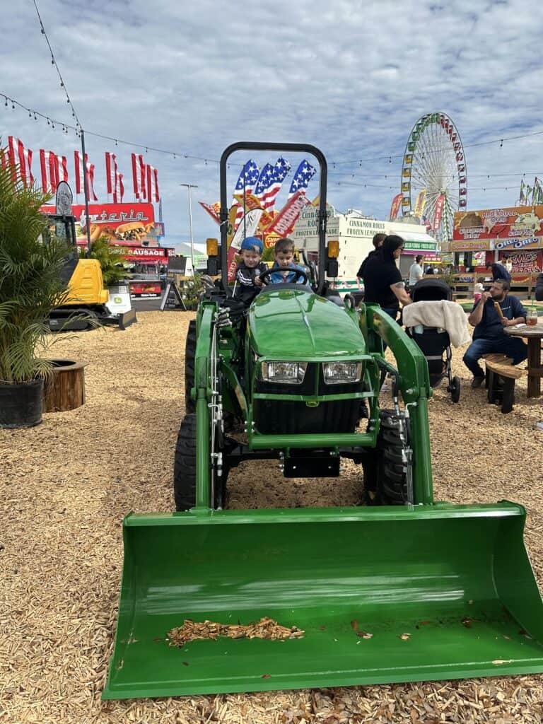 Two kids sitting on a green tractor at an outdoor fair, with a Ferris wheel and colorful signs in the background.
