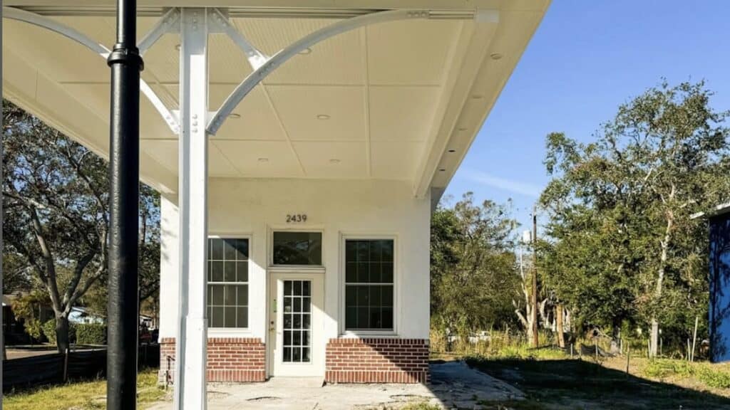 a small former fuel station. It has a brick base, and an off-white paint job. An shade structure is built over the front door/order window