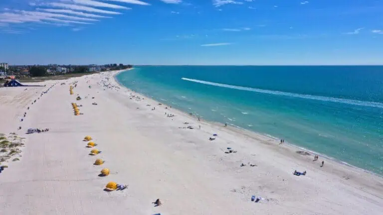 aerial view of a beach with lounge chairs and umbrellas visible. The water is a crystak blue, and the beach loungers have a yellow and blue color pattern against the white sand