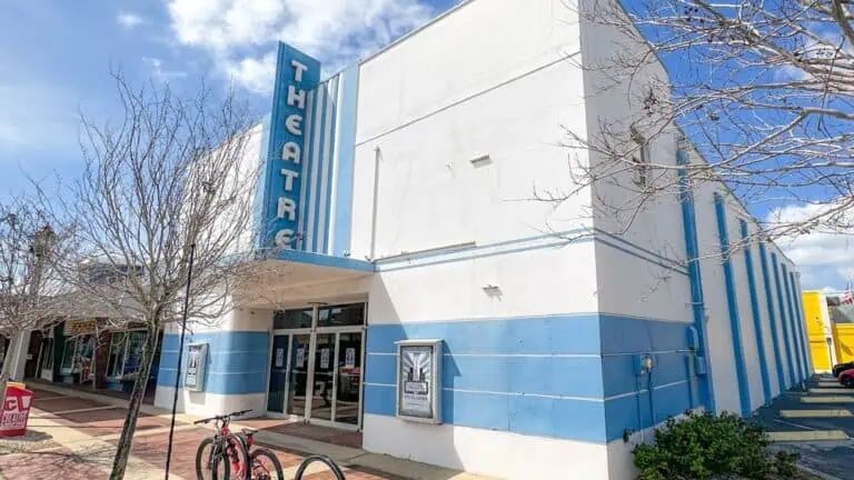 exterior of an old school movie theatre with a blue and white marquee. Trees surround the theatre on the sidewalk on a sunny day