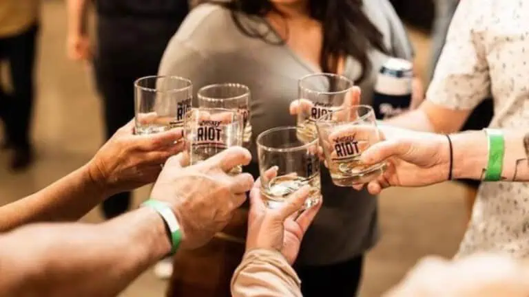 A group of people raising whiskey glasses labeled "Whiskey Riot" in a celebratory toast.