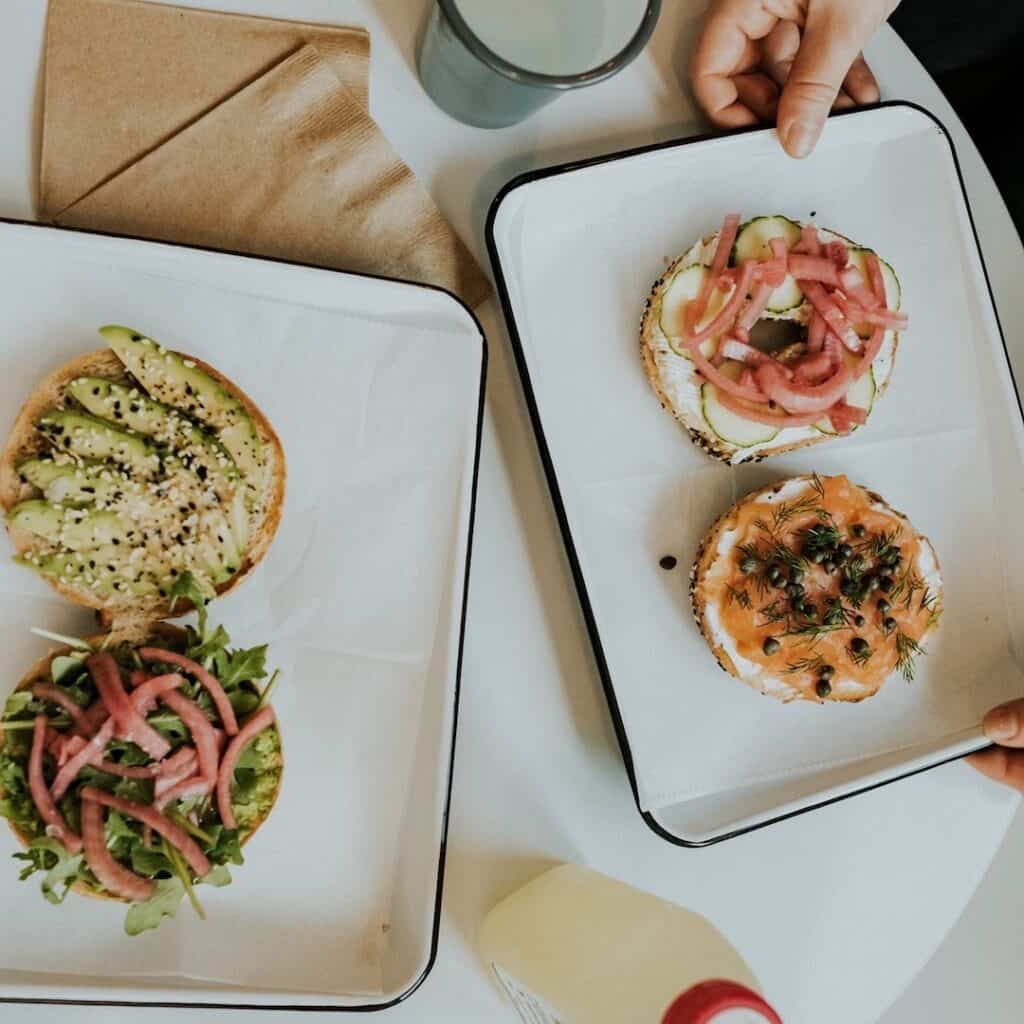 Two trays featuring open-faced bagels topped with avocado, pickled vegetables, smoked salmon, and herbs.