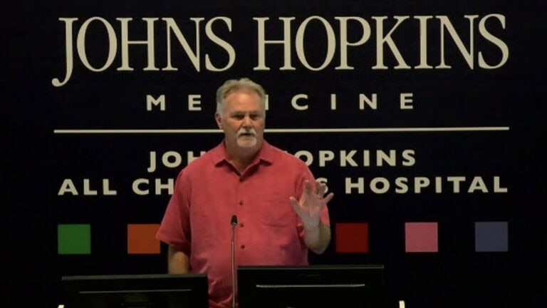 red-shirted man man speaking in front of a black background.