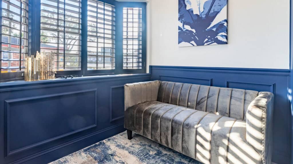 A cozy seating area with a gray velvet sofa, blue paneled walls, and natural light streaming through shutters.





