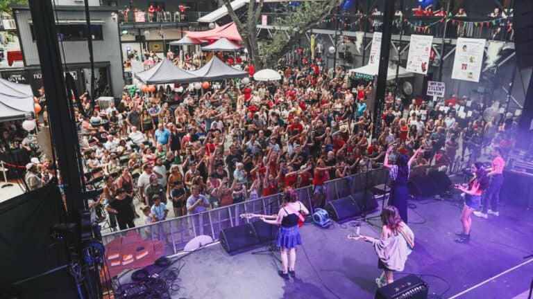 a group of performers on stage playing for a massive crowd. Purple lights cover the stage area. The venue is open air