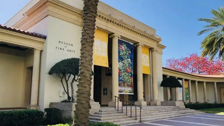 Exterior of a museum on a sunny day. Three colorful banners are draped over the front entrance. A tree with bright red blooms emerges from behind the museum. A brick walkway leads to the front door