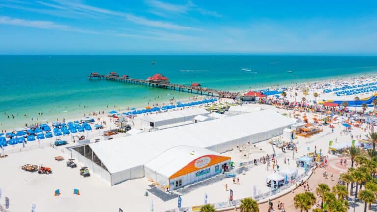 aerial view of a white sand beach with a large exhibit tent set up in front of royal blue cabanas. A long pier stretches out into the water. A pink sidewalk runs parrallel to the beach