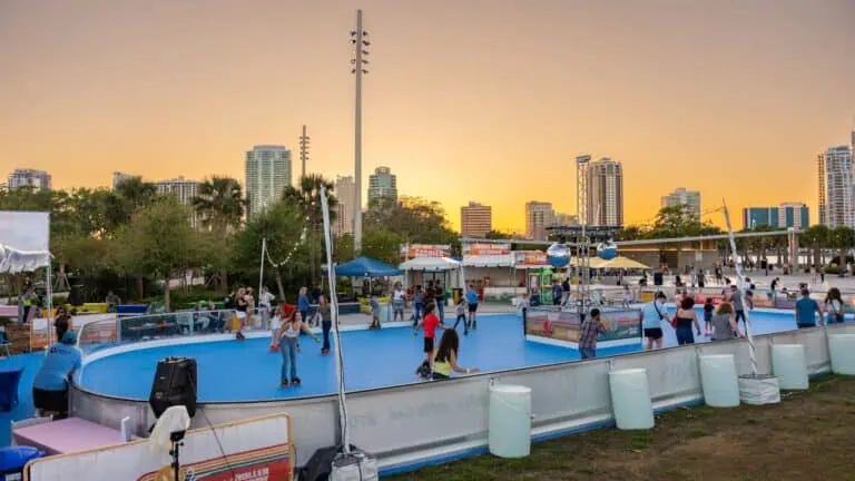 An outdoor roller rink at golden hour. The rink itself is blue and it's surrounded by multiple vendor tents. Two crystal balls hang over the center of the rink