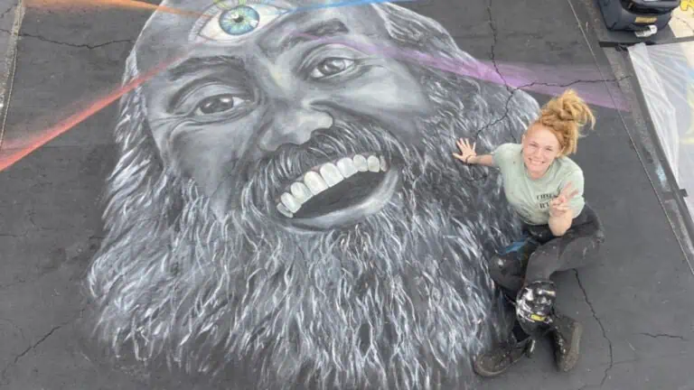 A woman is seated on the ground next to a vibrant chalk illustration of a bearded man, engaged with the drawing.