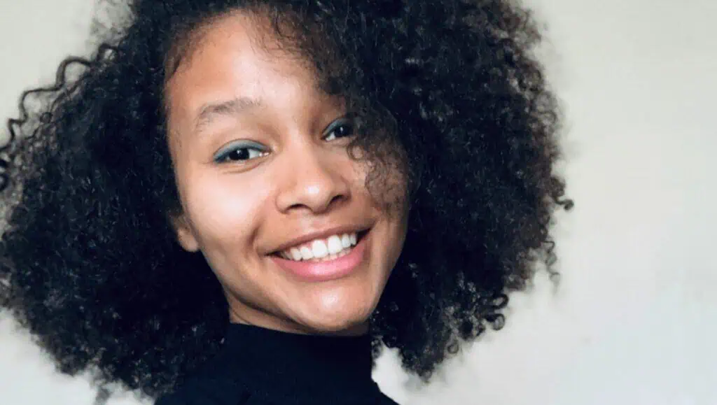 A portrait of artist Sam Modder. She's spiling and turned toward the camera. She has curly hair and is wearing a black turtleneck