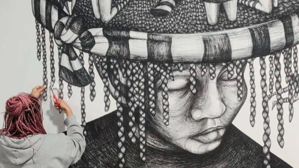 a black and white illustration by Sam Modder. The illustration is of a character with braided hair. A person stands near the illustration with a pair of scissors