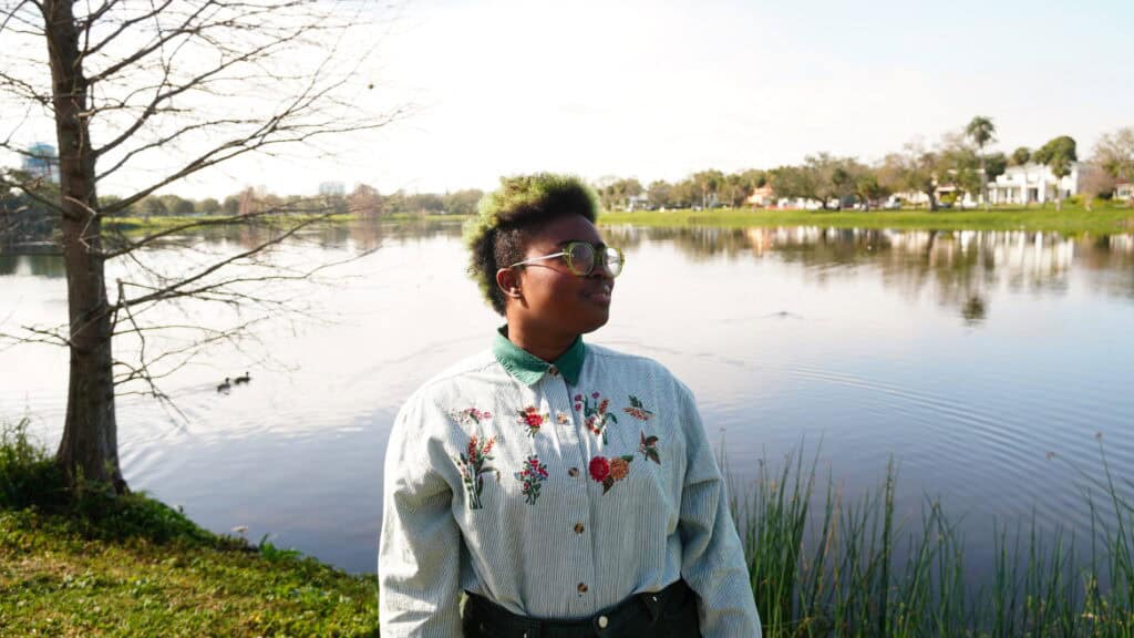 a therapist poses for a photo in a park. they are wearing a white button down shirt with flowers embroidered into it. They are standing in front of a lake on a sunny day.