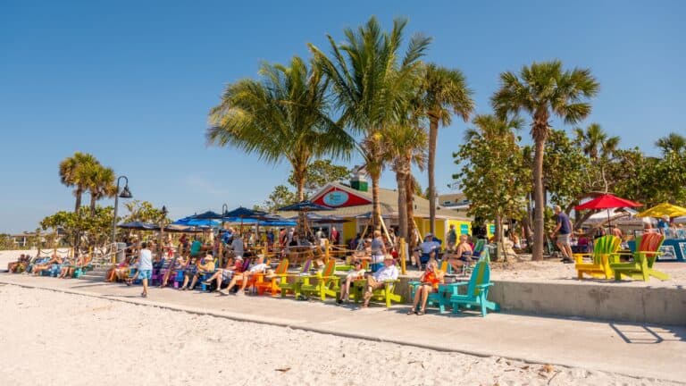 view from the sand of Paradise grille with palm trees and beachgoers in colorful plastic chairs out front on a sunny blue skies day
